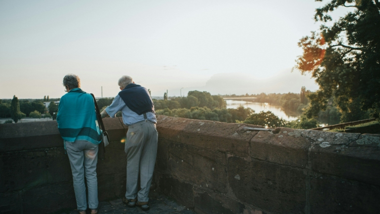 Zwei ältere Personen stehen an einer steinernen Brüstung und blicken auf eine malerische Flusslandschaft im Abendlicht.