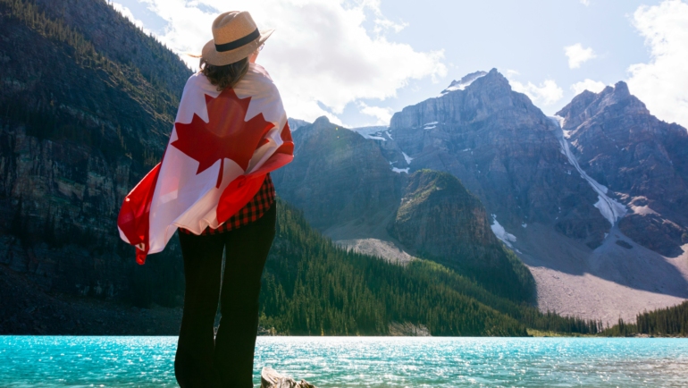 Eine Person steht mit dem Rücken zur Kamera am Ufer eines türkisfarbenen Sees, umhüllt von einer kanadischen Flagge. Sie trägt einen Hut und schaut in die Höhe zu einer beeindruckenden Berglandschaft.