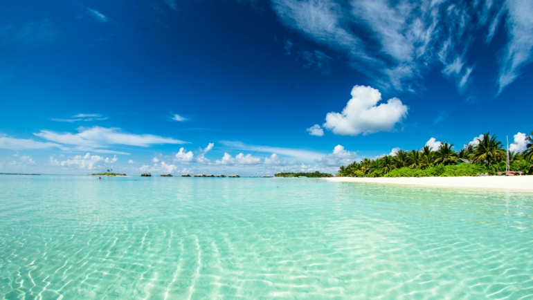 Ein friedlicher Strand mit klarem türkisfarbenem Wasser, feinem weißen Sand und Palmen unter einem klaren blauen Himmel mit ein paar weißen Wolken.