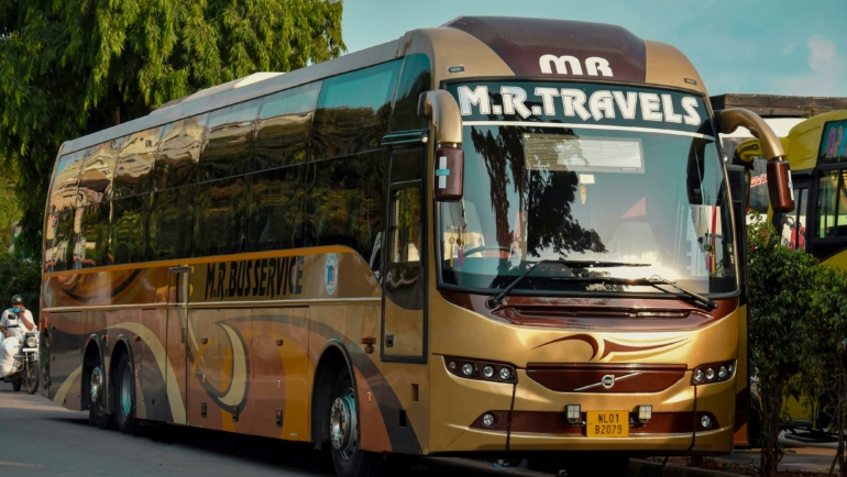 Ein großer, braun-goldener Reisebus mit der Aufschrift "M.R. Travels" steht am Straßenrand neben Bäumen.