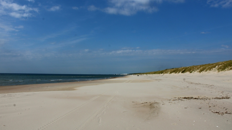 Ein weiter, menschenleerer Sandstrand mit sanften Dünen und Meer bei klarem, leicht bewölktem Himmel.