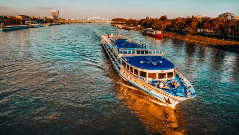 Ein weiß-blaues Schiff fährt auf einem breiten Fluss, umgeben von städtischen Gebäuden und herbstlichen Bäumen im Abendlicht.