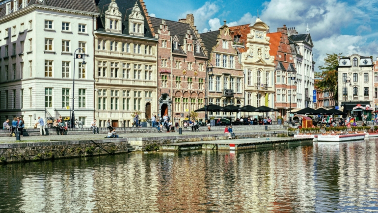 Historische Gebäude mit verzierten Fassaden spiegeln sich im Wasser eines Kanals. Menschen sitzen und flanieren am Ufer, während Boote am Rand liegen.