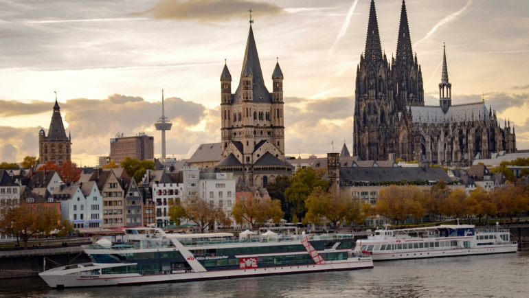 Der Kölner Dom überragt die Altstadt mit verschiedenen alten Häusern. Im Vordergrund zieht ein modernes Ausflugsschiff den Fluss entlang, während der Himmel von Wolken durchzogen ist.