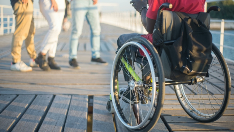 Eine Person im Rollstuhl sitzt auf einem Holzsteg und betrachtet ein Smartphone. Im Vordergrund sind drei unscharf abgebildete Personen zu sehen, die stehen und miteinander reden.