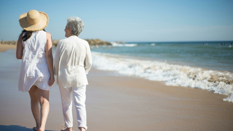 Zwei Personen gehen barfuß am Strand entlang, während die Wellen sanft an den Sand schlagen. Eine der Personen trägt einen Strohhut. Es ist ein sonniger Tag mit klarem Himmel.