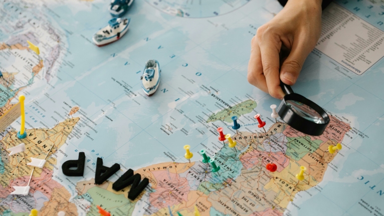 Eine Hand hält eine Lupe über eine Weltkarte, die mit bunten Stecknadeln markiert ist. Kleine Schiffsmodelle stehen ebenfalls auf der Karte.