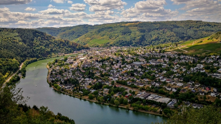 Die Aufnahme zeigt eine malerische Flusslandschaft mit einer Stadt, die sich entlang des Ufers schlängelt. Umgeben von grünen Hügeln und dicht bewaldeten Flächen breitet sich die Siedlung in einem Tal aus.
