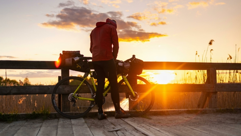 Ein Fahrradfahrer steht auf einer Holzbrücke bei Sonnenuntergang. Hinter ihm leuchtet die untergehende Sonne und taucht die Szene in ein warmes Licht.