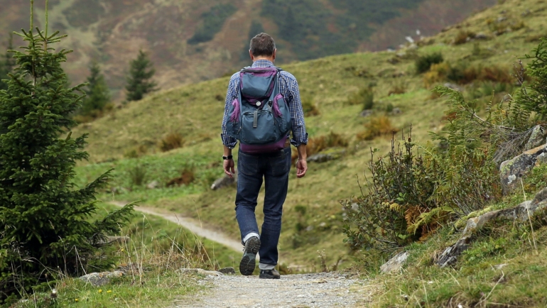 Ein Mann mit einem Rucksack wandert auf einem Pfad durch eine grüne, hügelige Landschaft.