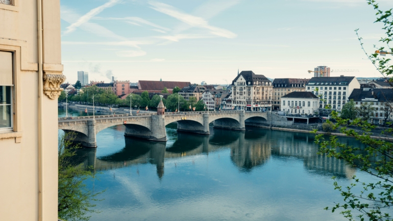 Eine historische Steinbrücke überspannt einen ruhigen Fluss, umgeben von malerischer Architektur und modernen Gebäuden im Hintergrund.