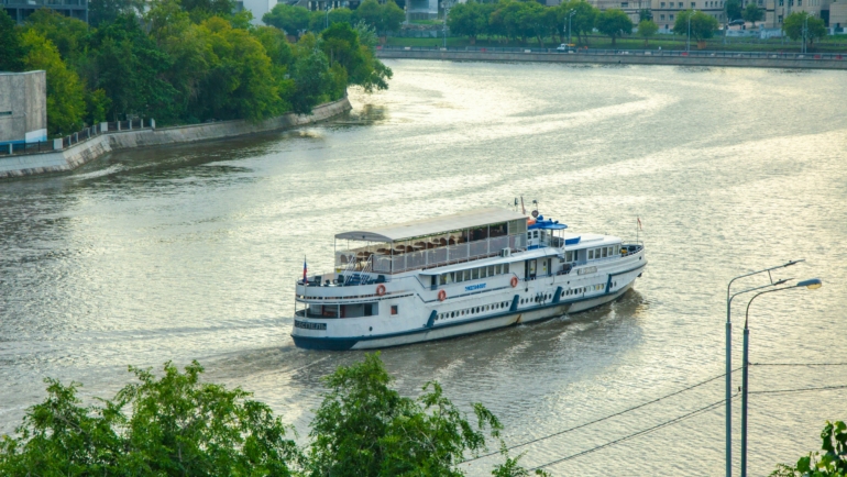 Ein weißes Schiff fährt auf einem breiten Fluss, umgeben von grüner Vegetation und bebauten Ufern. Im Hintergrund sind Gebäude zu sehen.