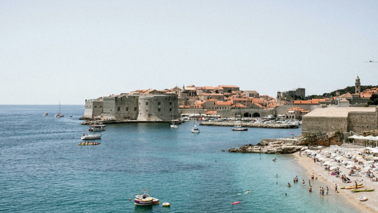 Blick auf eine Altstadt mit Stadtmauer am Meer und belebtem Strand im Vordergrund.