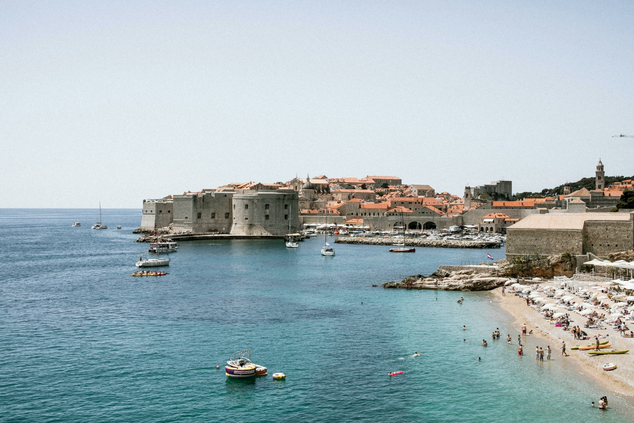 Blick auf eine Altstadt mit Stadtmauer am Meer und belebtem Strand im Vordergrund.